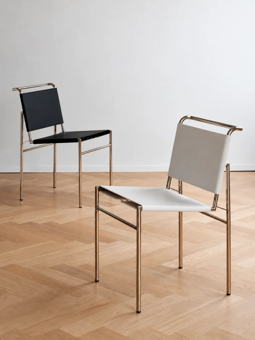 Two modern metal chairs with woven sling backs and seats, shown on a light wood herringbone floor against a white wall. One chair has a black seat and back, and the other has a white seat and back, both with gold-toned frames.