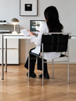 A woman sits at a desk in a bright, modern room, viewed from behind. She is wearing a white long-sleeve top and black trousers, seated on a black and silver chair.