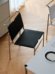 A modern chair with a black upholstered seat and backrest and a chrome metal frame, shown on a light wood herringbone floor in an interior setting.