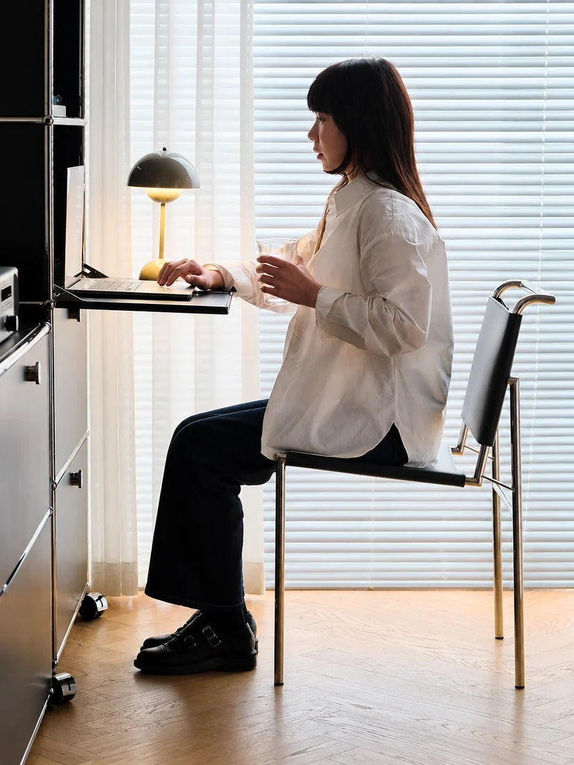 A woman sits in a modern office chair at a desk, wearing a white button-up shirt and dark trousers in a bright room with window blinds and a desk lamp.