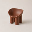 A small sculptural PE stool in a warm terracotta brown color, featuring a rounded, bowl-like seat supported by four cylindrical legs, set against a solid light beige background.