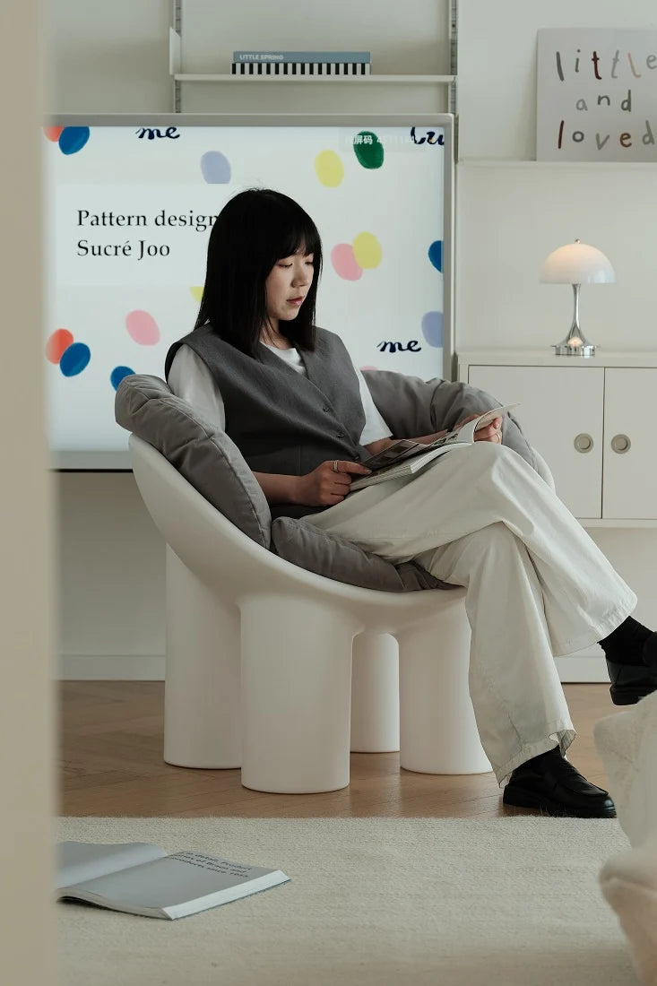 A woman sits in a white sculptural lounge chair in a bright, modern room, holding a magazine. A large white screen behind her displays the text 'Pattern design Sucre Joo' alongside colorful circular graphics.