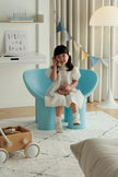 A young girl with dark hair sits smiling in a bright, modern room, wearing a white dress and white socks while sitting on a light blue molded chair. The room features a white desk, a floor lamp, and a light-colored rug.