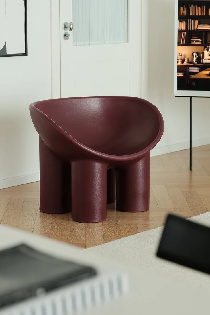 A modern sculptural armless chair in a deep burgundy color, set on a light wood floor in a minimalist room with a white door and wall art in the background.