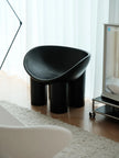 A modern black molded stool with three cylindrical legs, styled in a bright room with light wood floors, white curtains, and a white rug.
