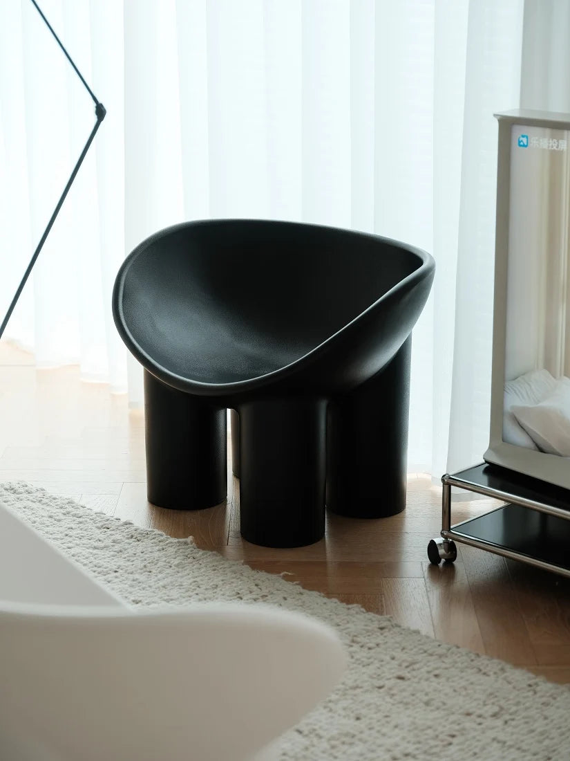 A modern black molded stool with three cylindrical legs, styled in a bright room with light wood floors, white curtains, and a white rug.