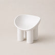 A minimalist white PE molded stool with four cylindrical legs and a rounded, asymmetrical seat, shown on a warm beige background.