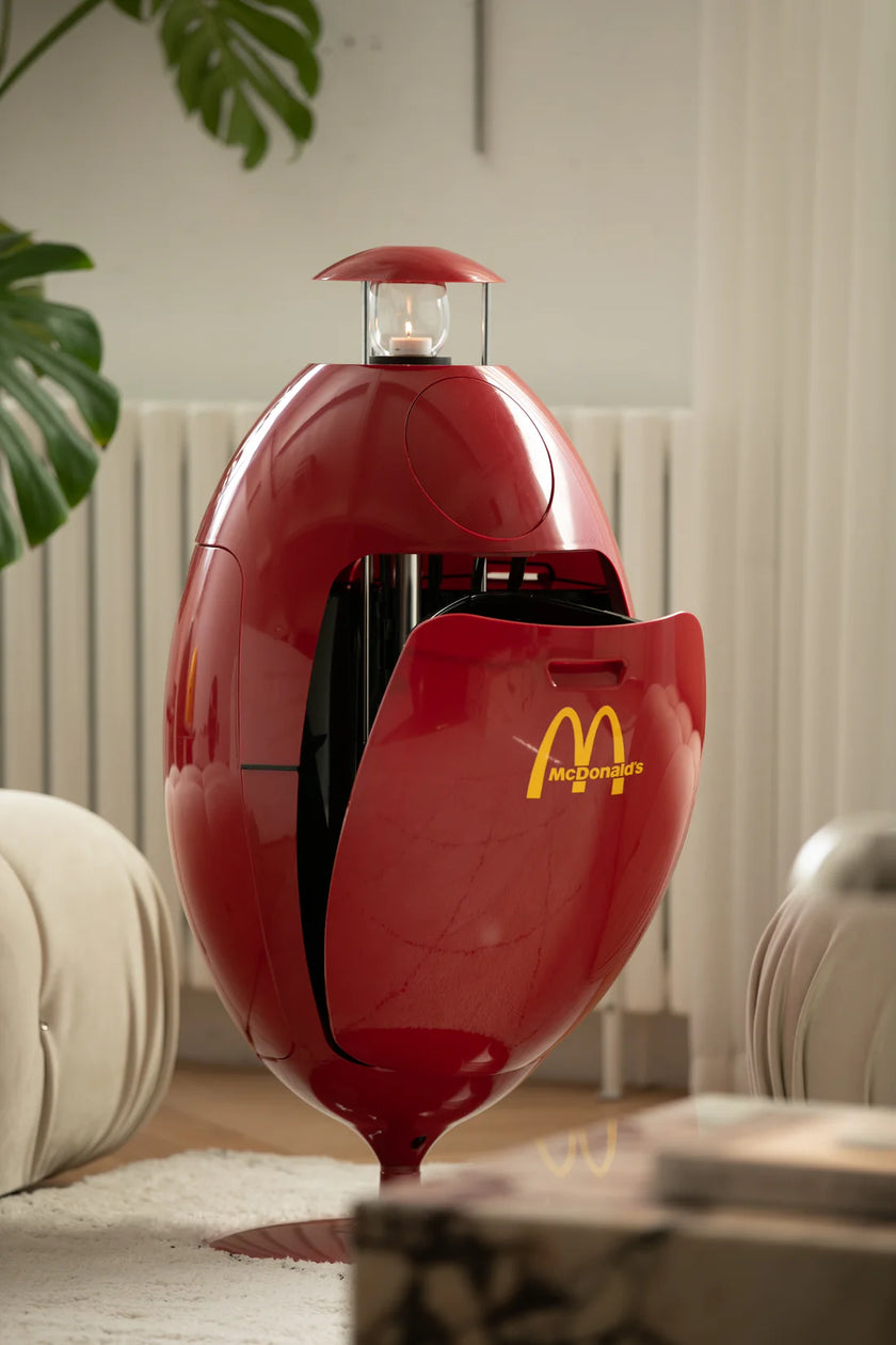 Red and black recycle bin with McDonald's logo on a table in a room with a plant and radiator.