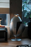 A modern egg-shaped floor lamp with a metallic silver finish and a small built-in light on top, shown in a stylishly decorated room with a window, plant, and side table.