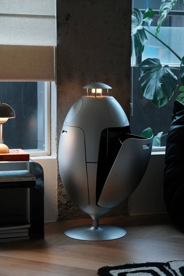 A modern egg-shaped floor lamp with a metallic silver finish and a small built-in light on top, shown in a stylishly decorated room with a window, plant, and side table.
