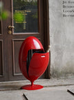 A glossy red egg-shaped outdoor trash can with a small bottle sticking out of the side, placed on a stone sidewalk in front of a brown door and textured wall.