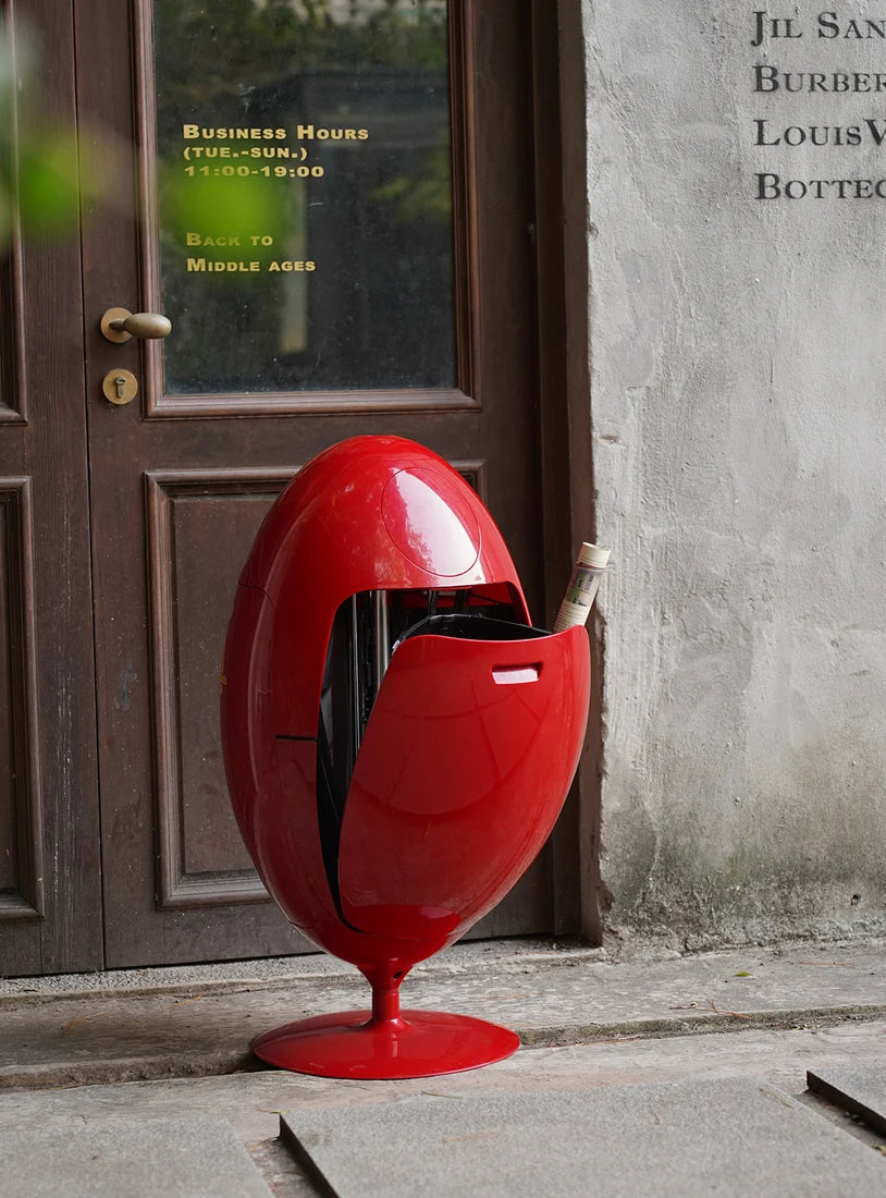A glossy red egg-shaped outdoor trash can with a small bottle sticking out of the side, placed on a stone sidewalk in front of a brown door and textured wall.