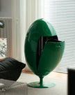 A glossy dark green egg-shaped pedestal table or cabinet with an open front panel, shown in a modern living room setting near window blinds.