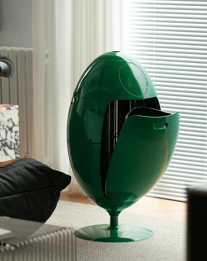 A glossy dark green egg-shaped pedestal table or cabinet with an open front panel, shown in a modern living room setting near window blinds.