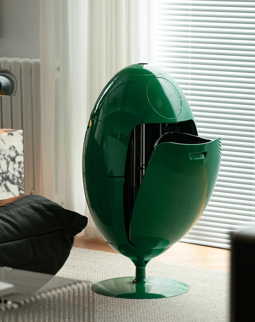 A glossy dark green egg-shaped pedestal table or cabinet with an open front panel, shown in a modern living room setting near window blinds.