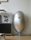 A metallic silver egg-shaped pedestal speaker on a light wood floor against a white wall, next to a wire shelving unit with books and a framed print.