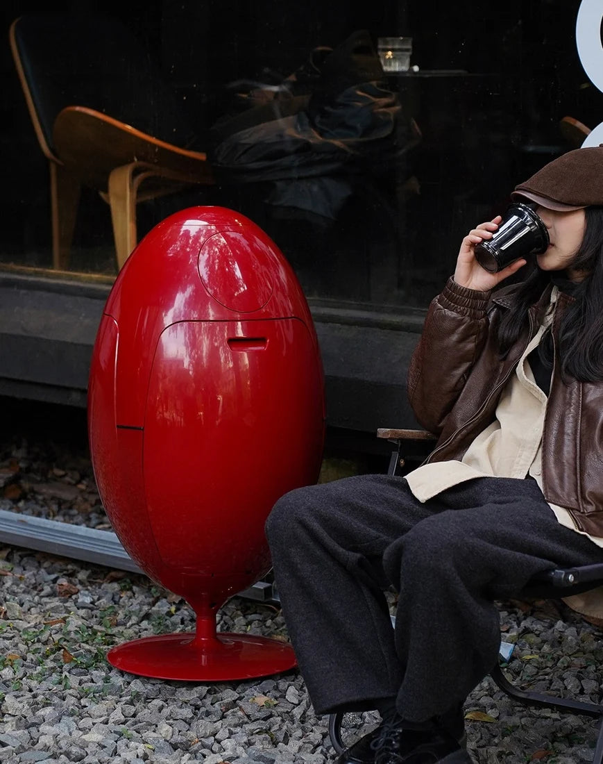 A person sitting on a chair outdoors next to a large glossy red egg-shaped object on a base, with gravel on the ground and a dark storefront in the background.