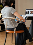 A woman sits in a modern chair with a curved chrome backrest and a brown leather seat, viewed from behind as she reads a book at a desk with a laptop in a home office setting.