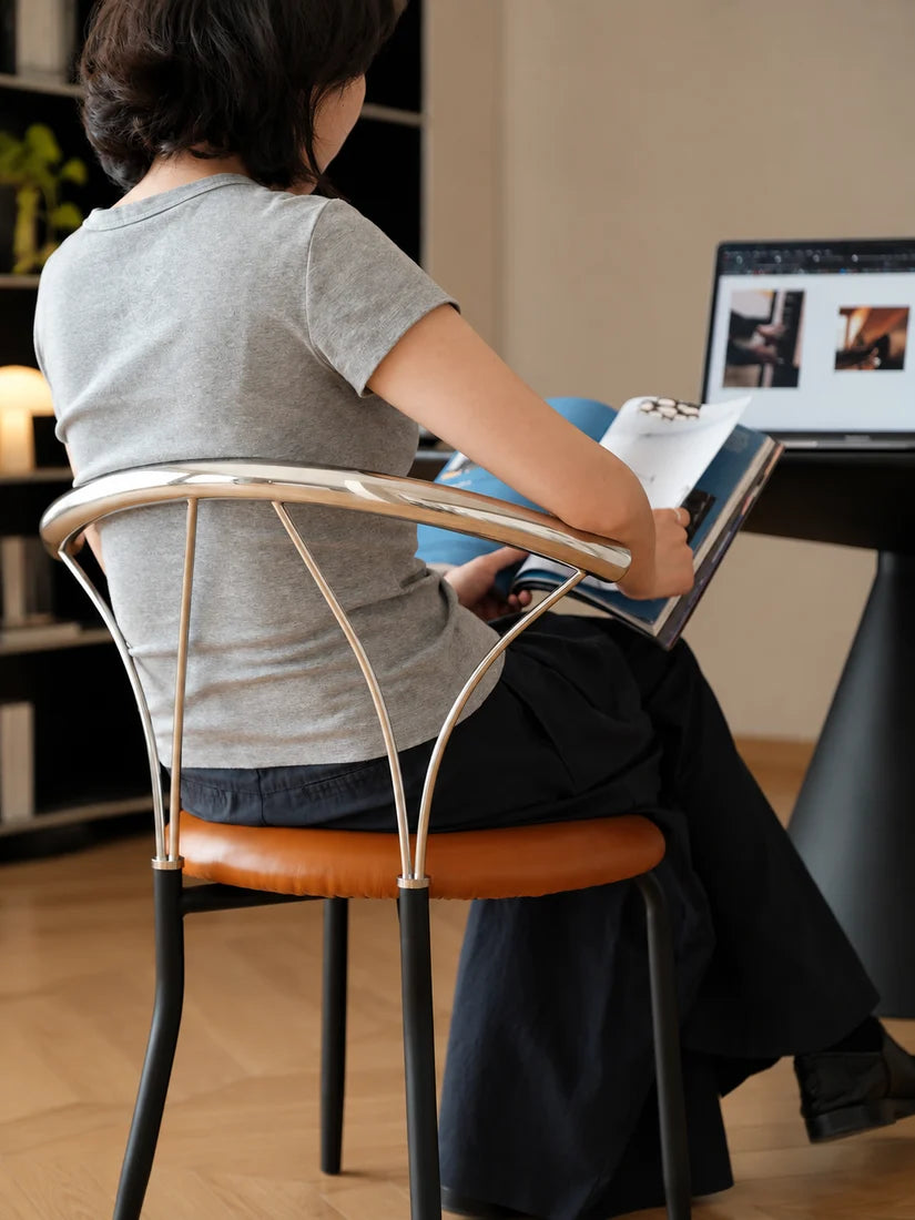 A woman sits in a modern chair with a curved chrome backrest and a brown leather seat, viewed from behind as she reads a book at a desk with a laptop in a home office setting.