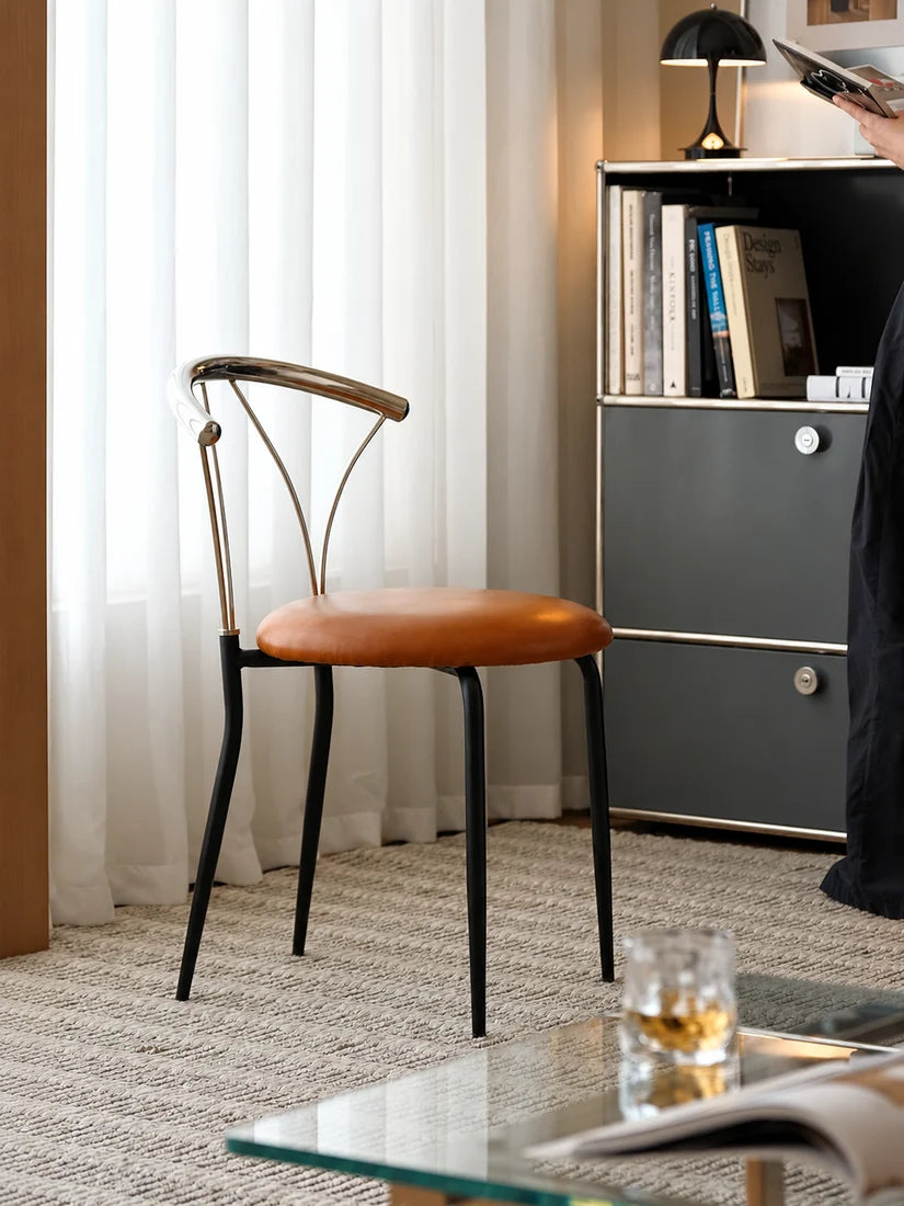 A mid-century modern accent chair with a curved backrest, black metal legs, and a brown leather seat cushion, set in a contemporary room next to a gray cabinet.