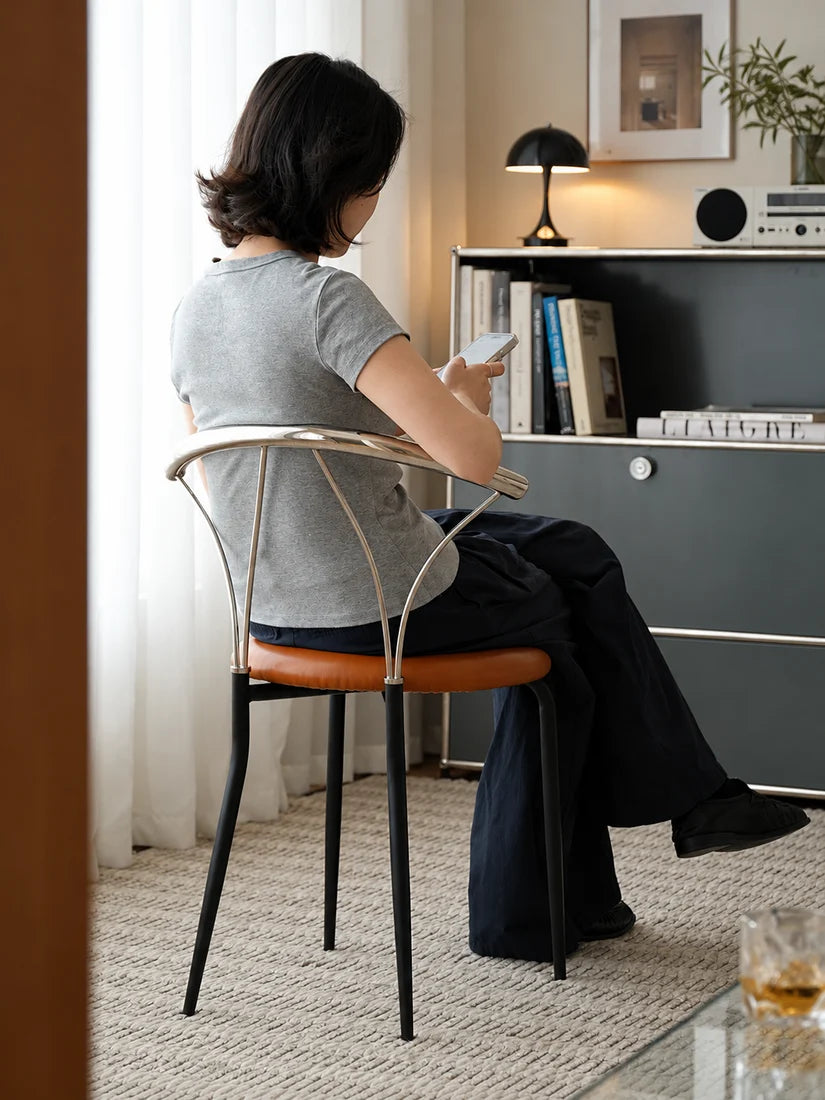 A woman sits on a modern metal chair with a curved backrest and a brown leather seat, viewed from behind as she looks at her phone in a stylishly decorated room.