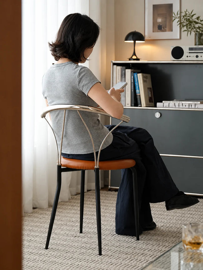 A woman sits on a modern metal chair with a curved backrest and a brown leather seat, viewed from behind as she looks at her phone in a stylishly decorated room.
