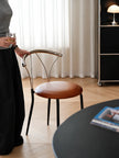 A woman moving a modern chair with a curved chrome backrest and a brown leather seat, viewed from behind as she reads a book at a desk with a laptop in a home office setting.