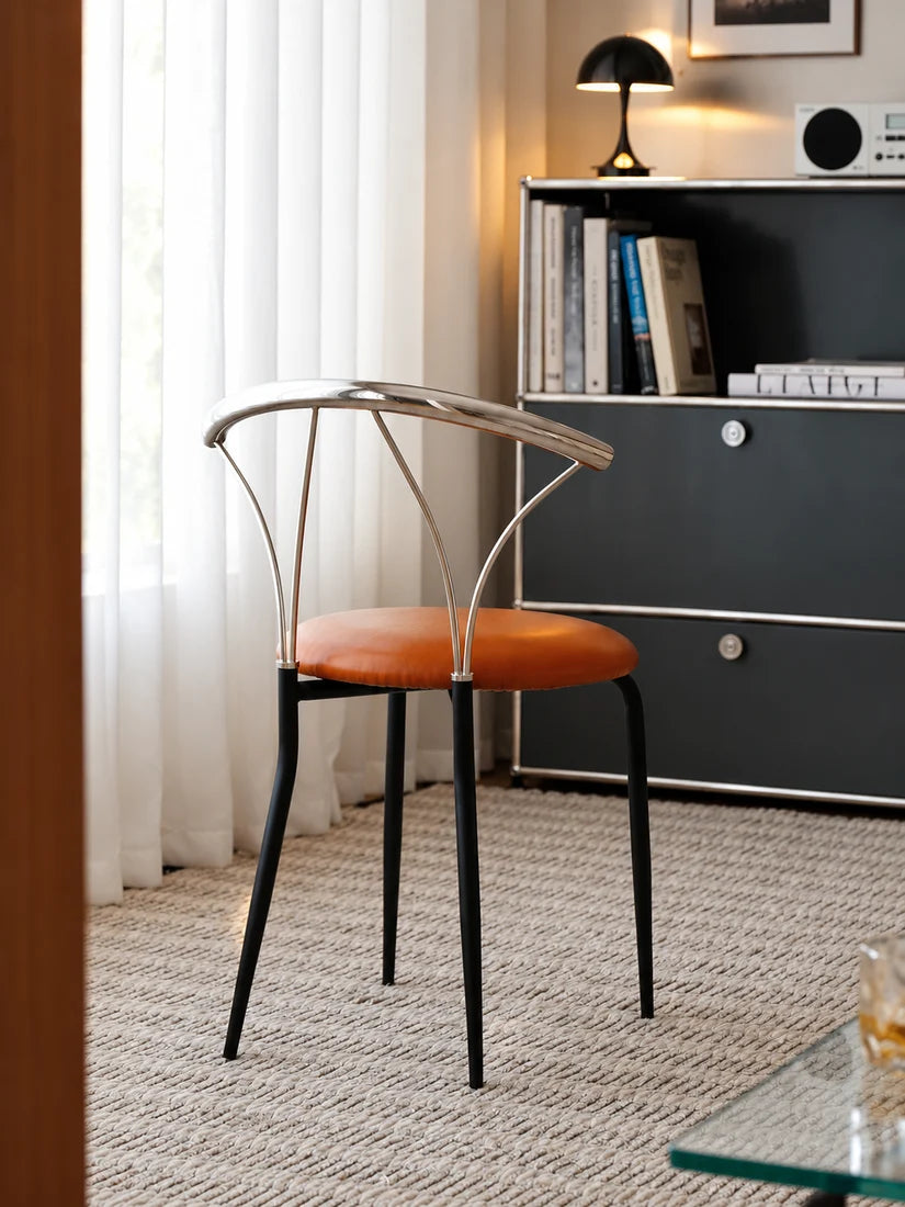 A modern dining chair with a curved silver metal frame, black legs, and an orange-brown upholstered seat, positioned in a contemporary room next to a dark gray cabinet.