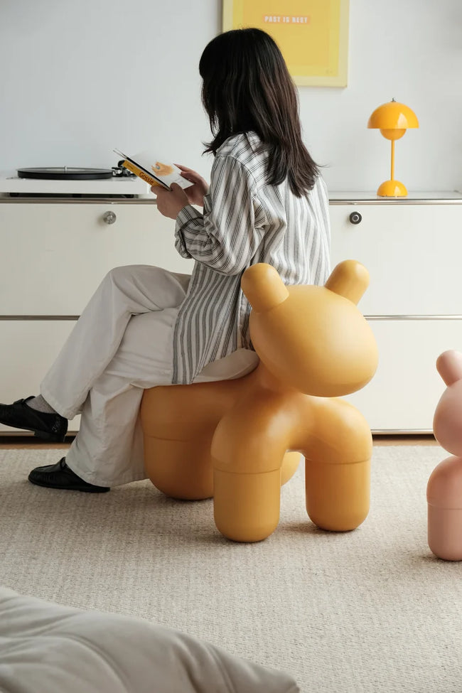 A woman sits on a mustard yellow bear-shaped children's stool in a bright, modern room, holding a tablet. A second pink bear stool is partially visible on the right.