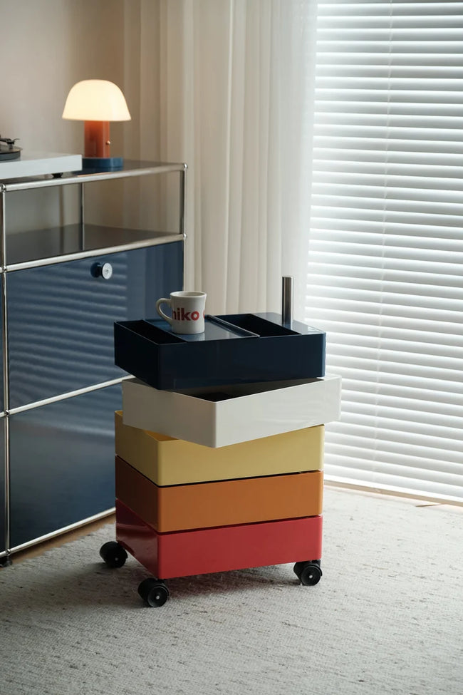 A rolling storage cart with stacked drawers in navy, white, yellow, orange, and red, shown in a bright room next to a blue cabinet and window blinds.
