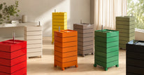A set of modern, multi-tiered rolling storage bins in various colors including red, orange, green, yellow, gray, beige, and black, arranged in a bright, modern room with natural light.