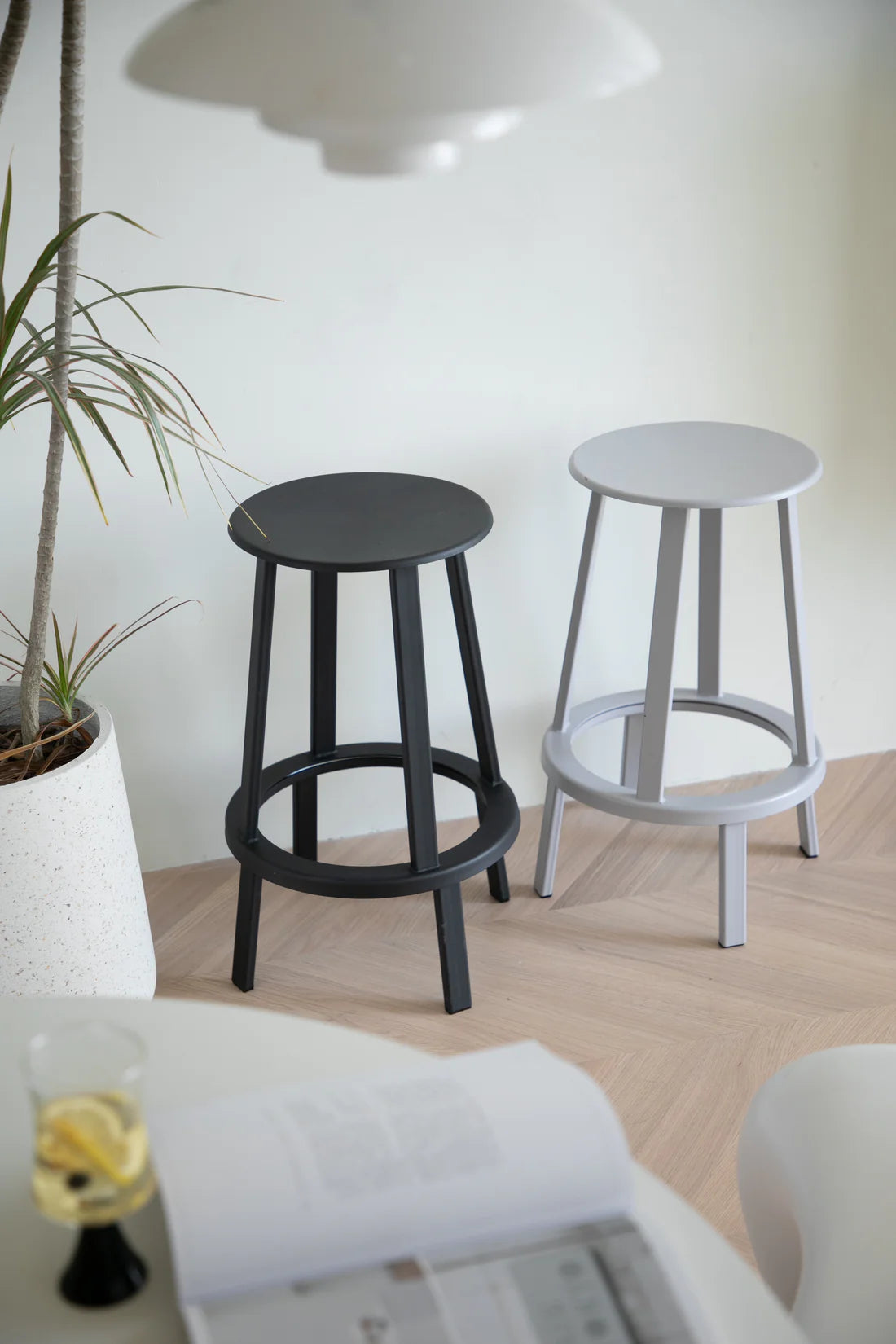 Two bar stools, one black and one silver, on a wooden floor with a plant and a glass of water in the background.