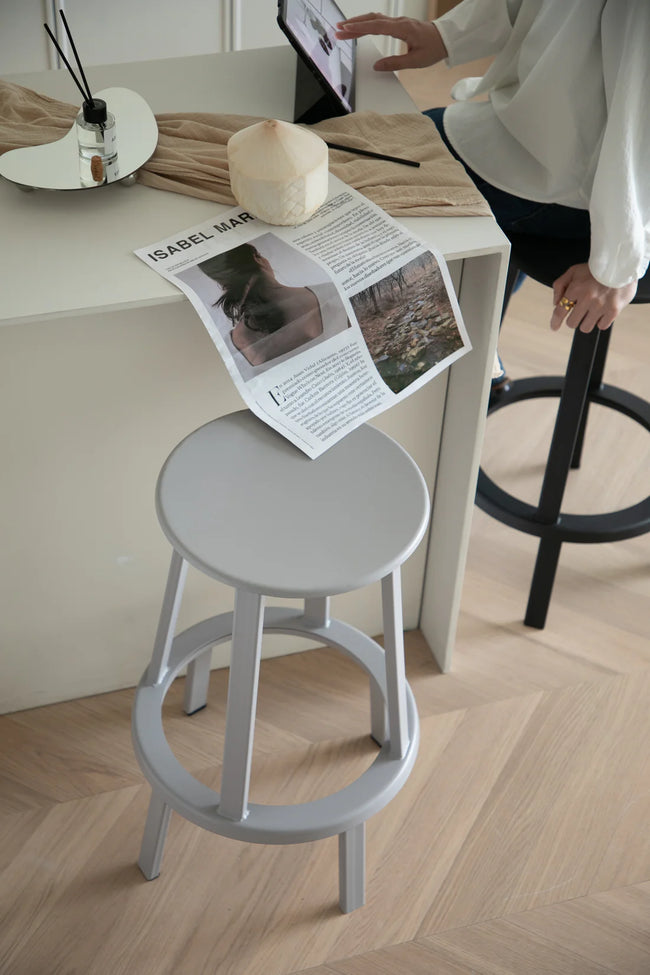 Gray metal counter stool with a magazine on a wooden floor, lady using a tablet in the background