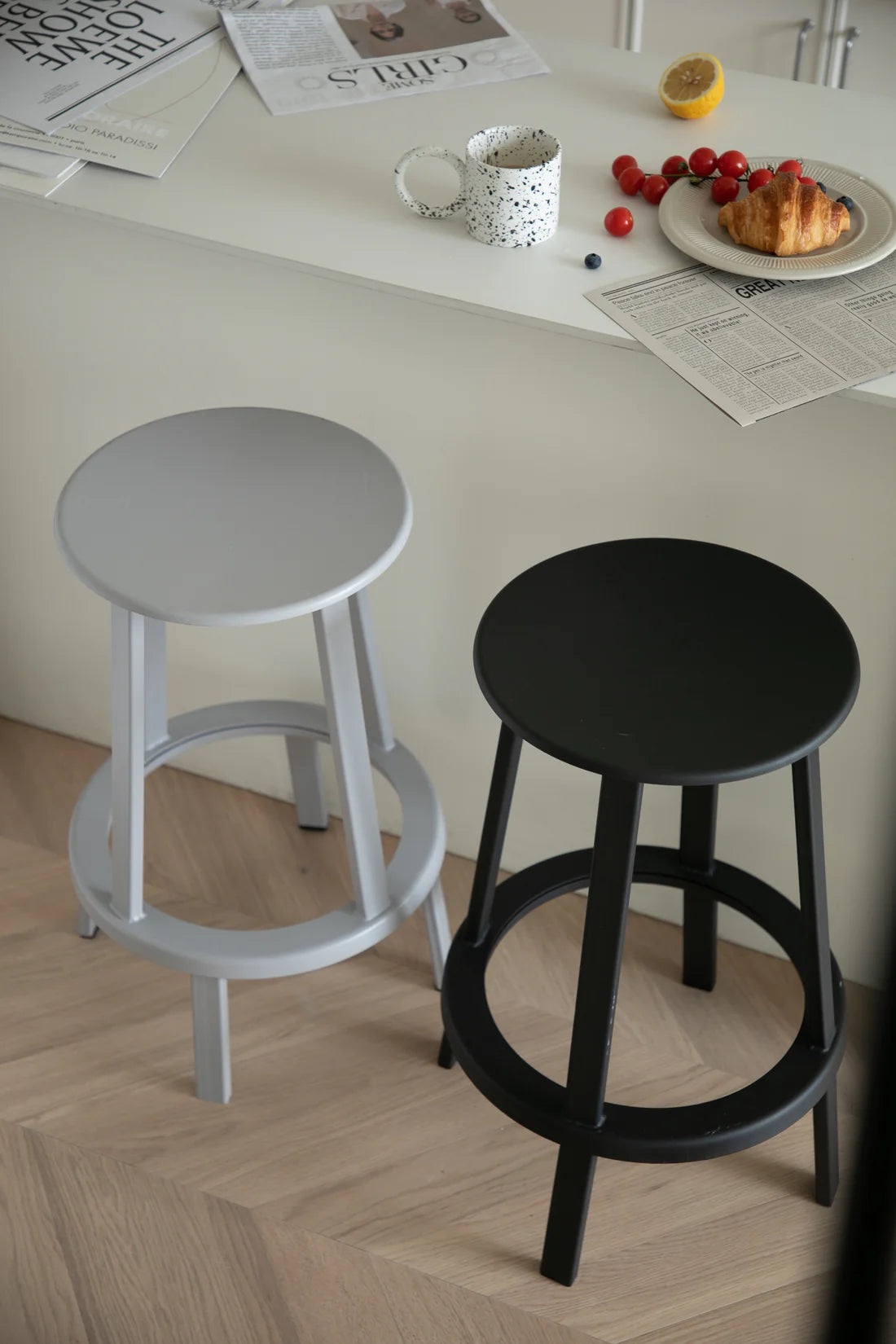Two bar stools, one gray and one black, on a wooden floor with a table and food items in the background.