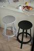 Two bar stools, one gray and one black, on a wooden floor with a table and food items in the background.