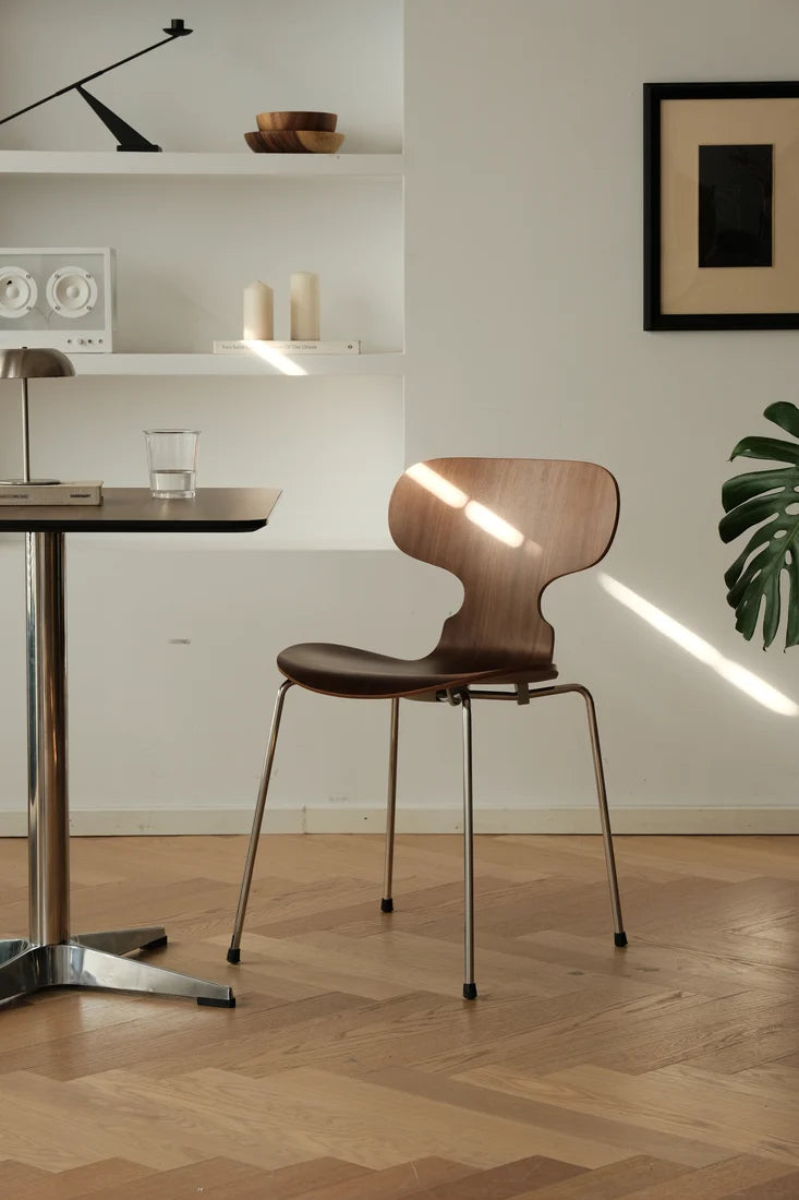 A modern wooden dining chair with a curved backrest and slim metal legs, positioned in a minimalist dining room setting with a table, shelves, and warm sunlight streaming through.