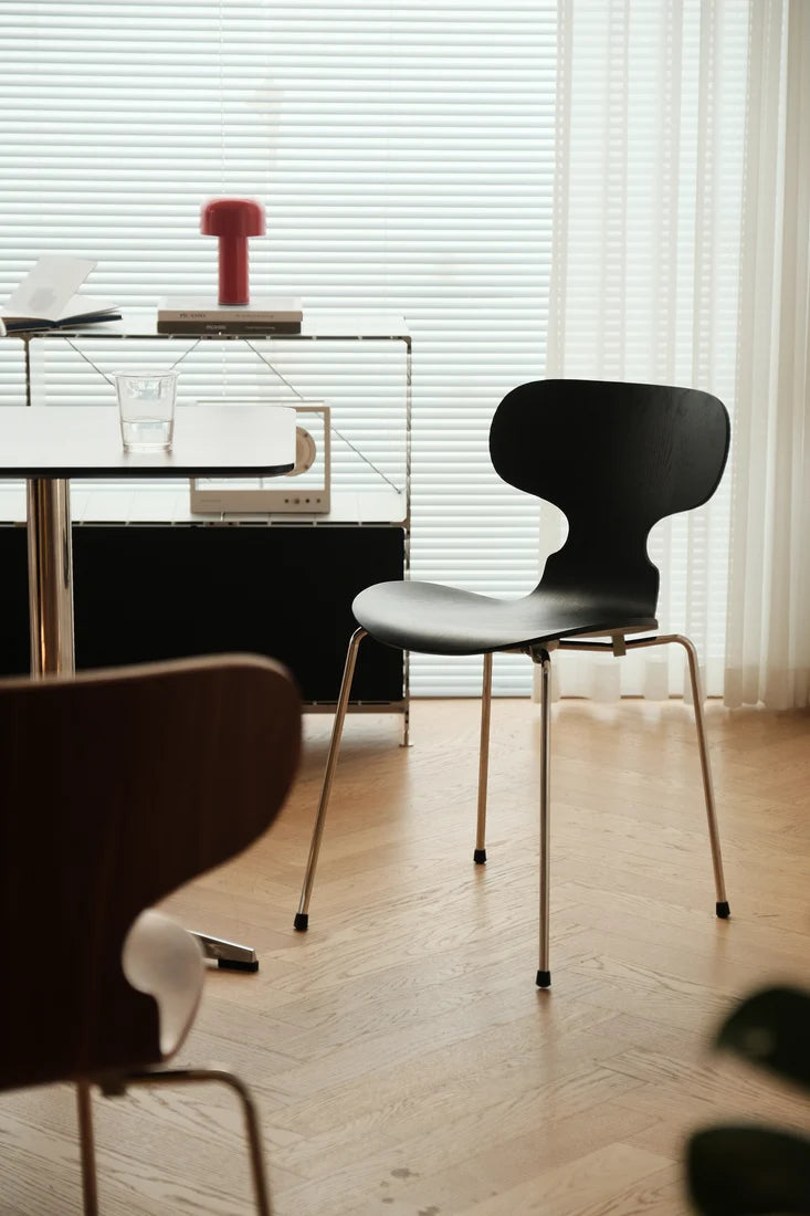 A modern black molded plastic dining chair with a curved backrest and slim gold metal legs, positioned in a bright, contemporary room with light wood floors and a glass side table in the background.