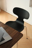 A modern black molded chair with a curved back and thin metal legs, positioned next to a dark wood desk with a laptop on a light wood floor.