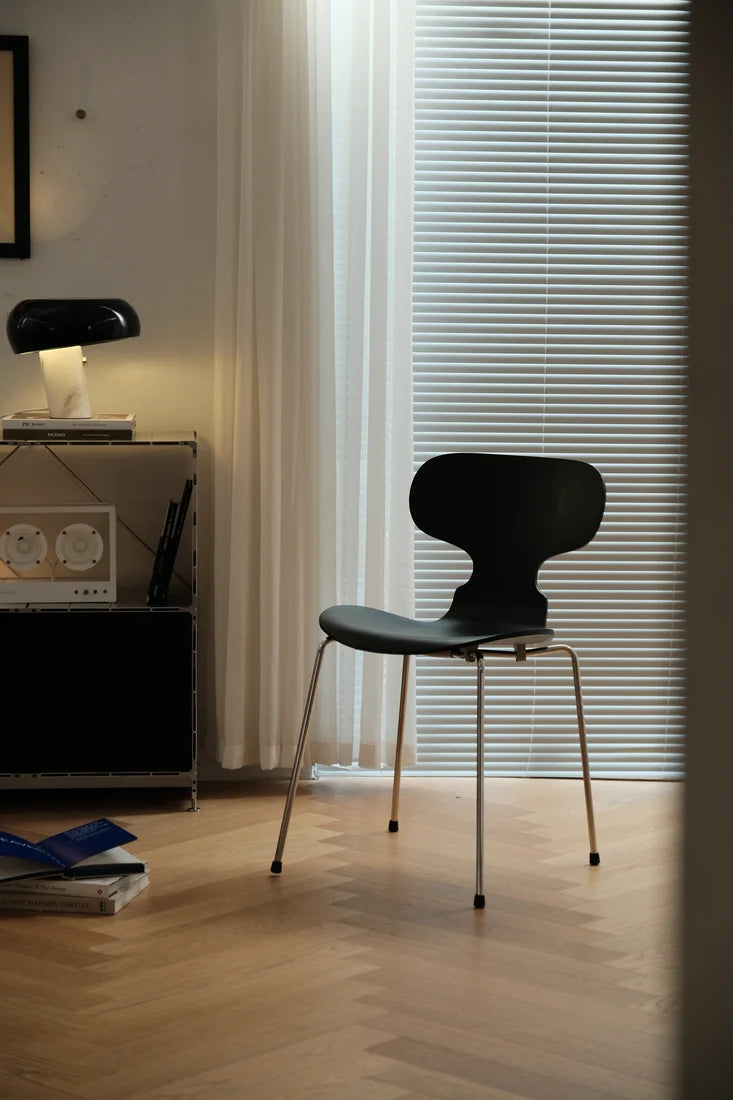A modern black molded plastic chair with slim metal legs, positioned in a minimalist room with light wood floors, white curtains, and window blinds.