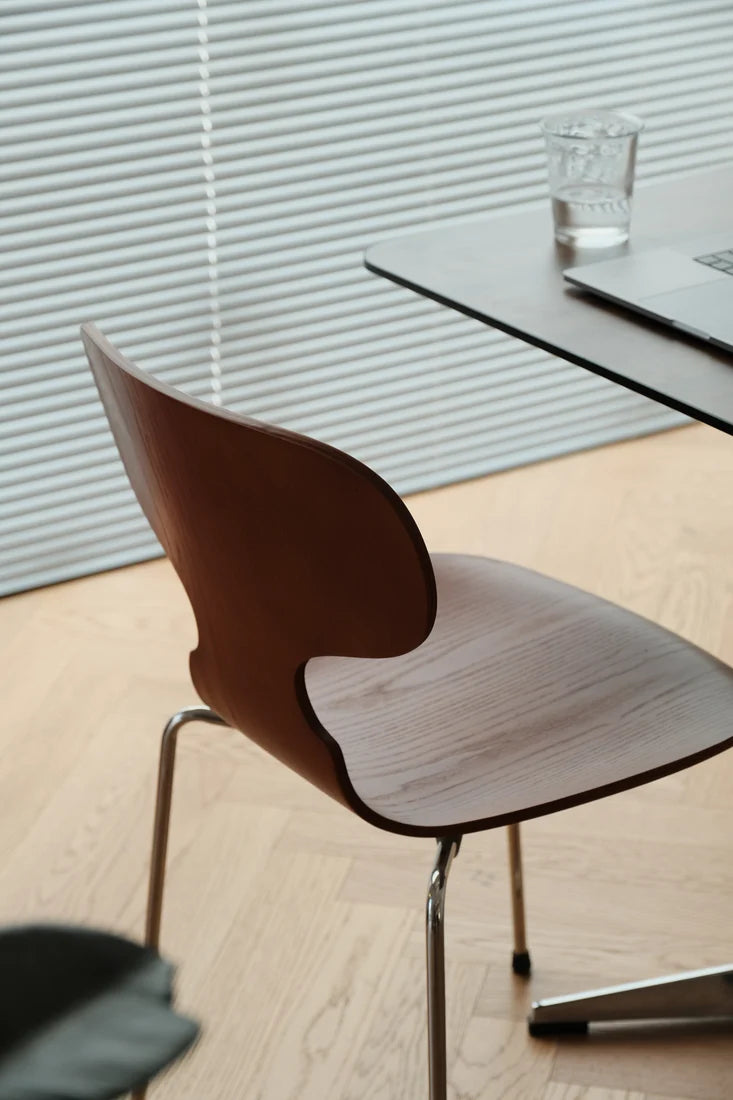 A modern wooden dining chair with a curved backrest and light wood seat, positioned next to a dark table in a bright room with light wood flooring and window blinds.