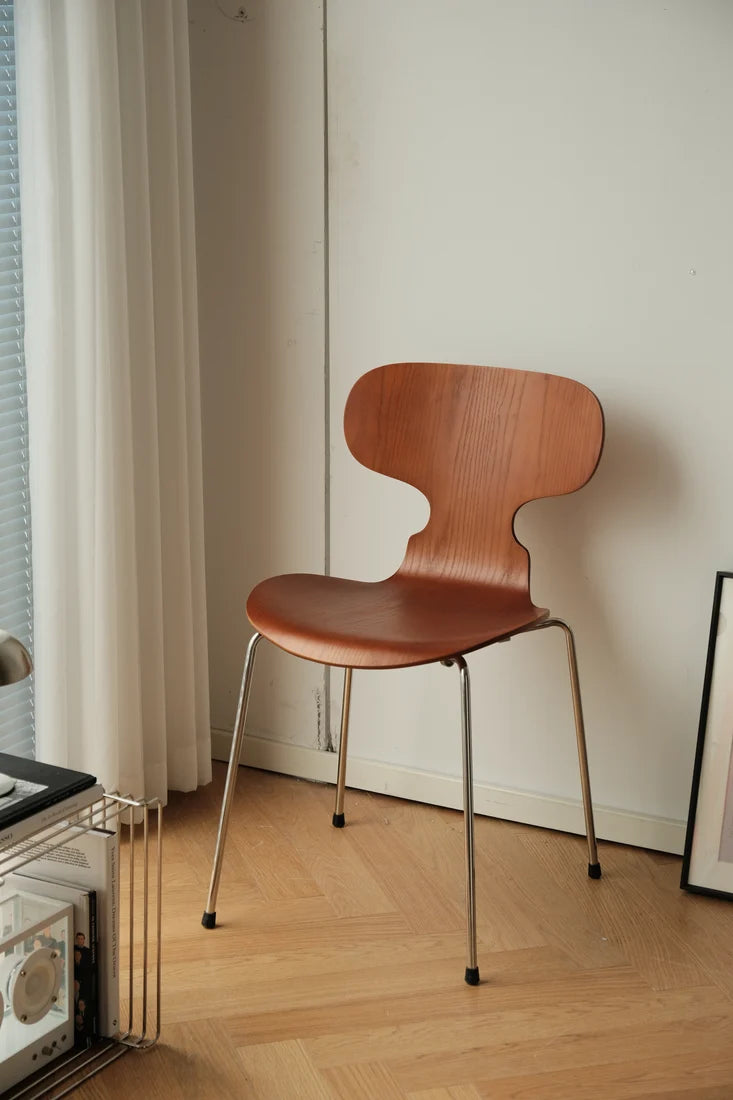 A mid-century modern wooden dining chair with a curved backrest and slim metal legs, positioned in a minimalist room with light wood flooring, a white wall, and a curtain.