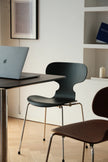 A modern black molded plastic chair with thin metal legs sits at a dark table with a laptop on a light wood floor, with another brown chair partially visible in the foreground.