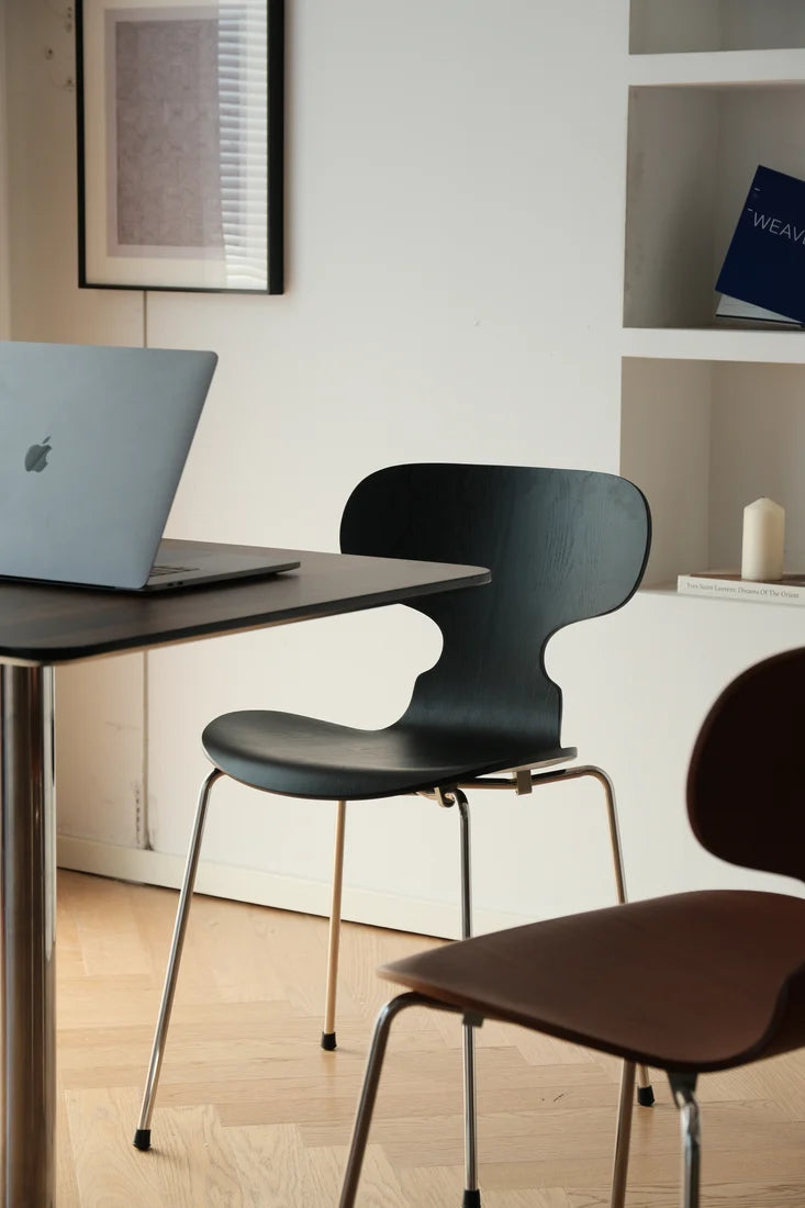A modern black molded plastic chair with thin metal legs sits at a dark table with a laptop on a light wood floor, with another brown chair partially visible in the foreground.
