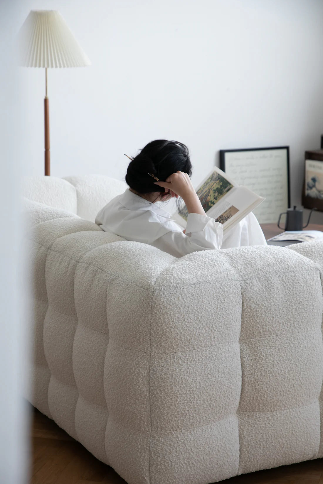 Women sitting on a white sofa reading a book in a room with a lamp and framed picture.