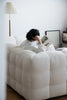 Women sitting on a white sofa reading a book in a room with a lamp and framed picture.
