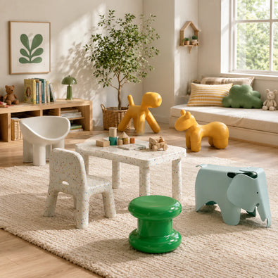 A styled children's playroom scene with a small white speckled table, two matching chairs, a green pedestal stool, a light blue elephant stool, and animal-shaped toys in a bright room with a window, rug, and bookshelf.