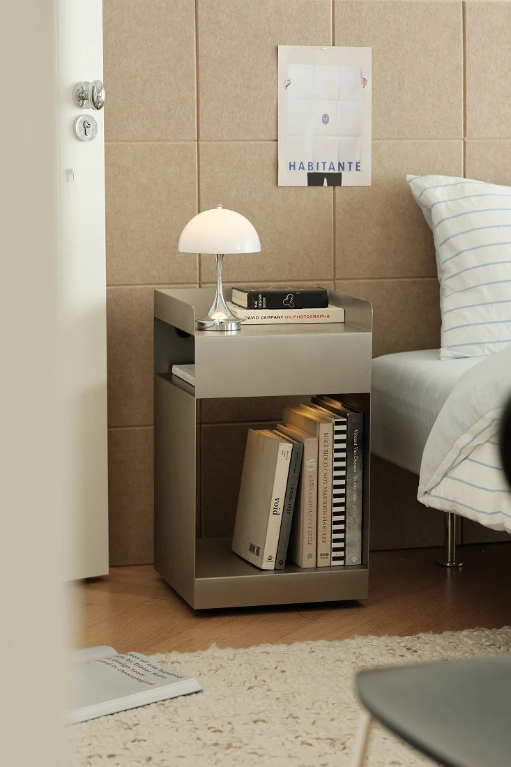 A modern metallic gray bedside table with an open shelf, styled in a bedroom setting next to a bed with a striped pillow and a white table lamp on top.