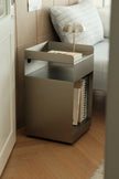 A modern metallic side table with a tray top, open lower shelf, and small lamp and books on top, placed in a real indoor room with wood flooring and a sofa in the background.