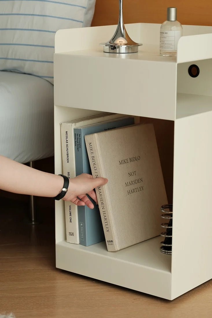A modern off-white bedside table or organizer shown in a bedroom setting, holding books and a person's hand is reaching toward them. A lamp and bottle sit on the top surface, and part of a bed is visible in the background.