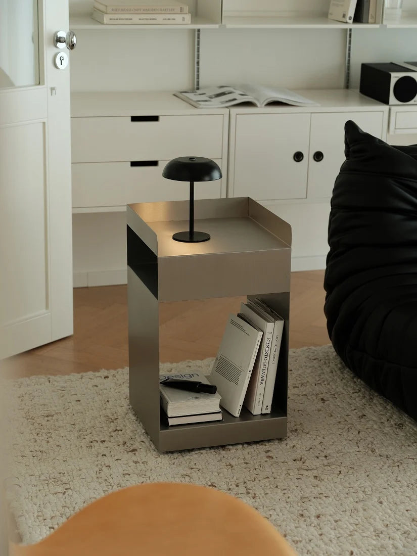 A modern square side table with a brushed silver metal finish and black accents, styled in a minimalist room with a black floor lamp on top, books in the open shelf, and a black beanbag chair nearby.